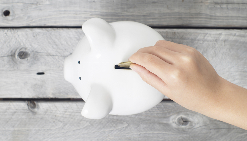 Asian boy hand dropping a coin into white piggy bank against wooden grey background
